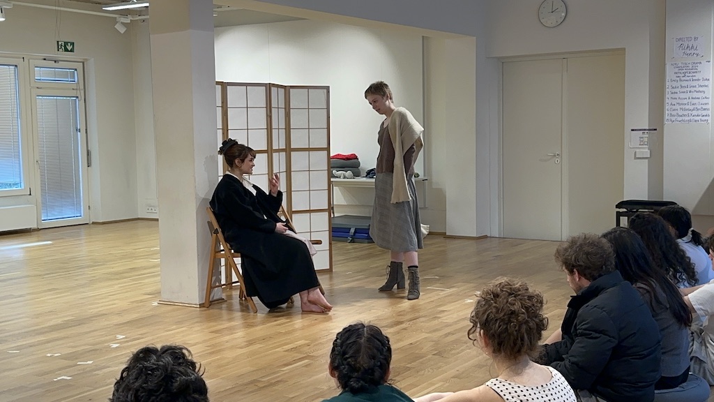Two actors, one seated and one standing, are in mid-performance of 'Faith Hope Charity'. A few students sit in the audience watching the performance.